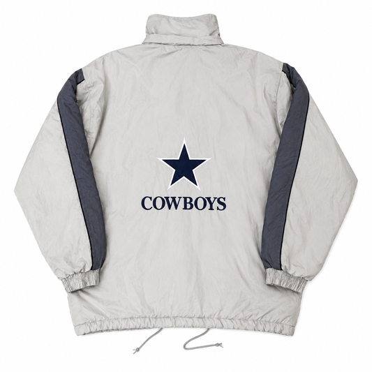 A light tan puffer jacket shown from the back, featuring a navy blue Dallas Cowboys star logo and the word 'COWBOYS' printed in the center, with dark gray sleeve stripes.
