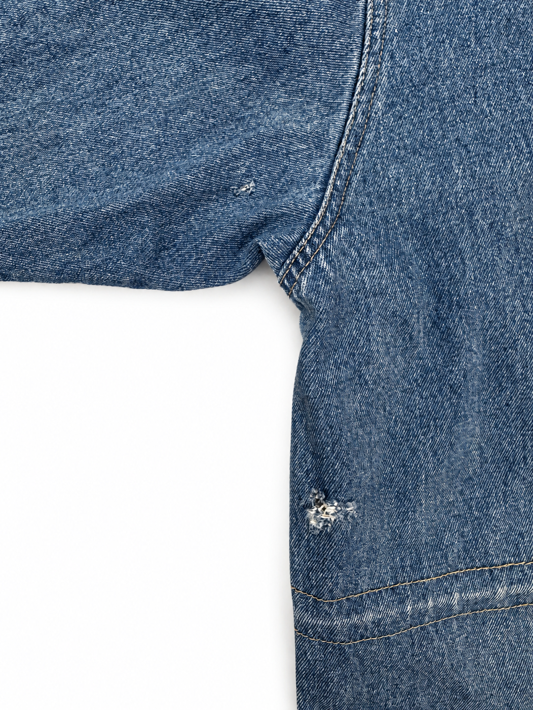 Close-up of a blue denim garment, showing the side seam, hem, and a small distressed detail on a white background.
