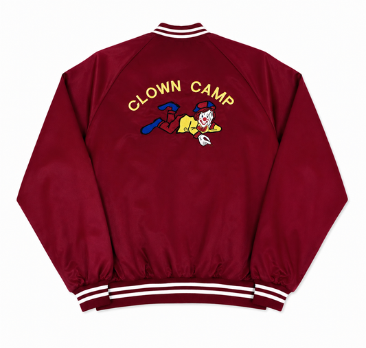 A red satin varsity jacket shown from the back, featuring 'CLOWN CAMP' embroidered in yellow above a cartoon clown graphic, with white and navy striped ribbed collar, cuffs, and hem.