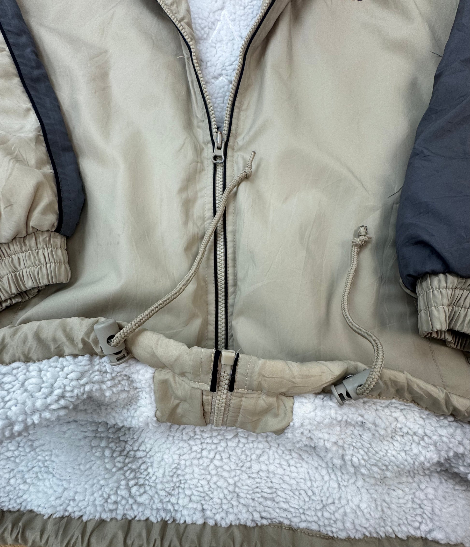 Close-up of a tan zip-front jacket with a white sherpa lining, elastic cuffs, and dark gray sleeve panels.