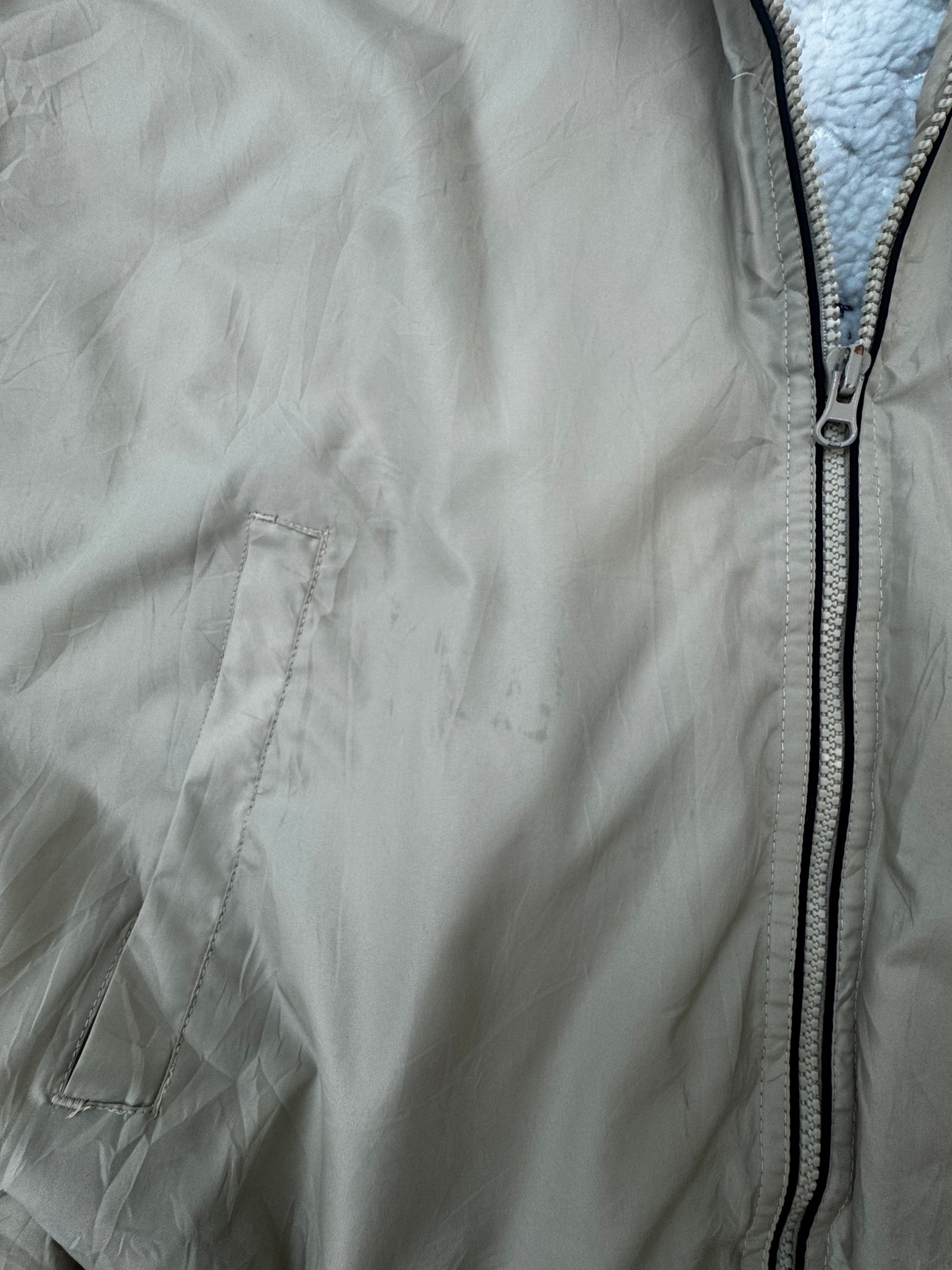 Close-up of a light tan lightweight jacket with a front zipper, a zippered chest pocket, and a light blue sherpa lining visible at the collar.